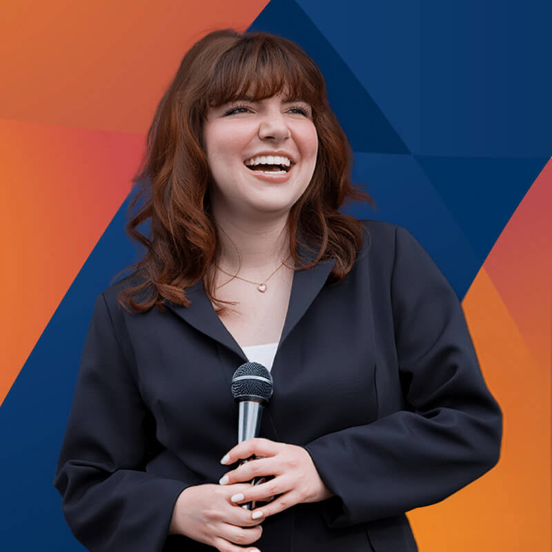 Woman with shoulder-length red hair holding a microphone and smiling, standing against an orange and blue background.